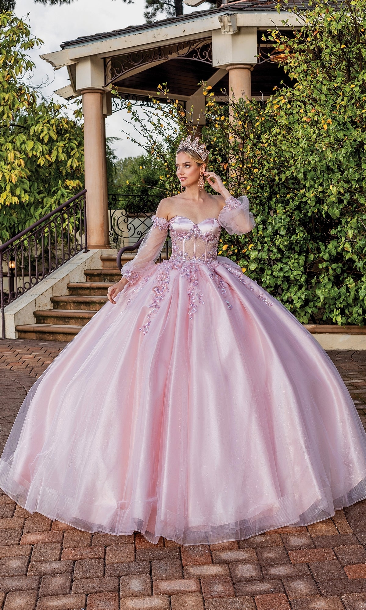 A woman stands outdoors on a brick path in the Quinceanera Dress 1824 By Dancing Queen, a blush pink ball gown with floral details and a crown, near stairs and greenery.